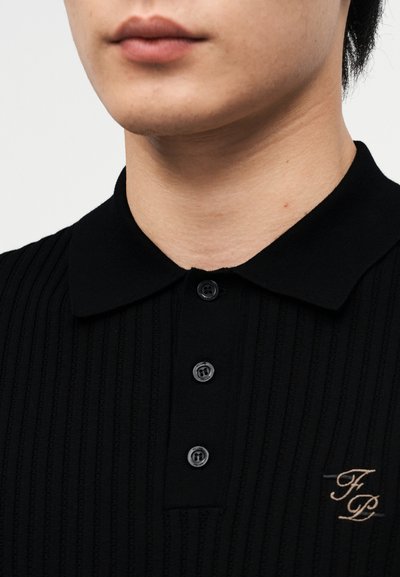 Κοντινό πλάνο κάτω μέρους προσώπου και λαιμού με μαύρο ριμπ πουλόβερ πόλο με τρία κουμπιά και μπεζ κεντημένα αρχικά "FP" στο στήθος.