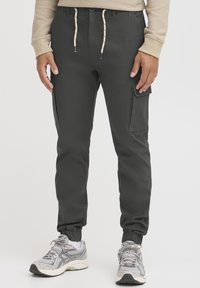 Cargo-byxor i mörkgrått tyg med elastiskt midjeband, uppvikta anklar, sidofickor och beige snören. Bärs med grå sneakers.