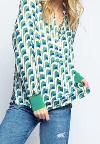 Blusa de manga larga con un patrón geométrico de ondas en azul y verde sobre un fondo blanco, con puños a rayas verdes.