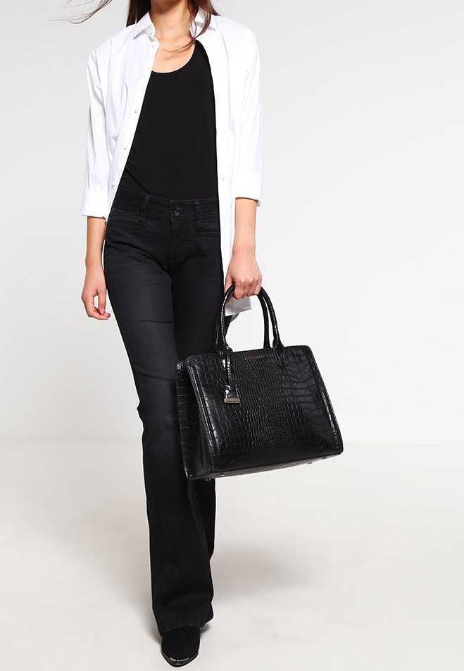 Femme portant une chemise blanche et un top noir avec un pantalon noir, tenant un grand sac à main noir texturé tout en marchant vers l'avant.