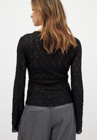 Black lace long-sleeve top with a fitted design, featuring a textured floral pattern. Worn with gray high-waisted trousers.