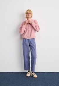 Persona con capelli biondi corti che regola una giacca in pile rosa, indossando pantaloni blu larghi e sneakers beige, in piedi contro un muro bianco e un tappeto blu.