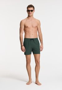 Zelené plavky s elastickým pásom a šnúrkou. Látka vyzerá hladko a je ľahká. Model s okuliarmi na slnko a bosý.