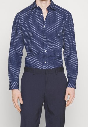 Mann trägt eng anliegendes dunkelblaues Hemd mit weißem Rautenmuster und dunkle marineblaue Hose, steht vor einfarbigem Hintergrund.