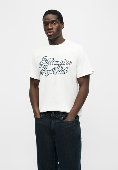 Νεαρός άνδρας που φοράει λευκό μπλουζάκι «Billionaire Boys Club» και σκούρο τζιν, στέκεται με τα χέρια στις τσέπες μπροστά σε απλό φόντο.