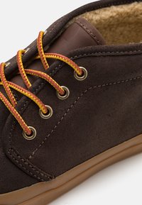 Brun mockaskor med gula och röda skosnören, rundad form, guldfärgade öljetter och en brun läderdetalj. Mjukt foder synligt inuti.