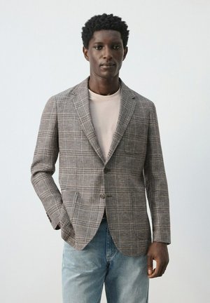 Muškarac obučen u sivi karirani blejzer preko bež majice i svijetlo plavih traperica, stoji ispred jednobojne svijetlo sive pozadine.