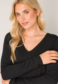 Black long-sleeve top with a V-neck design, featuring soft texture. Model has blonde hair styled in loose waves, wearing a gold necklace.
