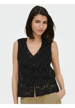 Femme aux cheveux foncés mi-longs, portant un top noir en crochet sans manches et un pantalon beige, une main dans la poche.