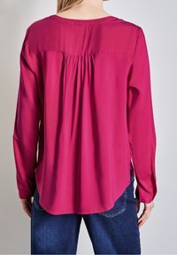 Long-sleeve blouse in fuchsia with a gathered pleat at the back, smooth fabric, rounded hem, and side slit details. Paired with denim.