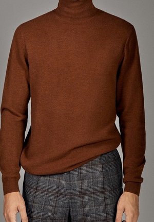 Mand iført en brun rullekravepullover og gråternet bukser, stående med armene afslappet langs siderne mod en ensfarvet baggrund.