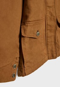 Poche de veste marron avec rabat sécurisé par un bouton-pression rond en métal et deux boutons-pression métalliques supplémentaires sur le bord inférieur.