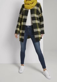 Kabát s velkým kostkovaným vzorem v černé a žluté barvě z vlněné směsi, spárovaný s bílým tílkem a tmavě modrými džínami, doplněný bílými teniskami a žlutým šálou.