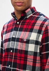 Hombre vestido con una camisa de cuadros en rojo, negro, blanco y gris, con botones, bolsillo en el pecho y logotipo bordado en el lado izquierdo.