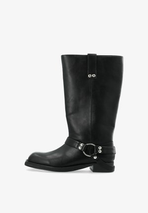 Bottes en cuir noir jusqu'au genou avec une forme large, une sangle avec un accent métallique circulaire, et un talon bas avec une semelle texturée.