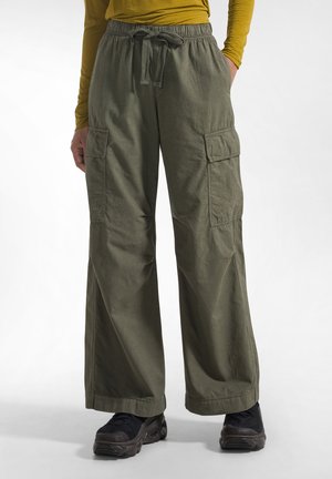 Person iført olivengrønne bukser med vide ben og sidelommer, sorte sneakers og en sennepsfarvet langærmet skjorte med den ene hånd i lommen.