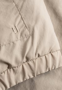 Béžová prešívaná bunda z látky s textúrovaným vzorom, so zberaným švom na leme a jemným logom vyšitým svetlou niťou.