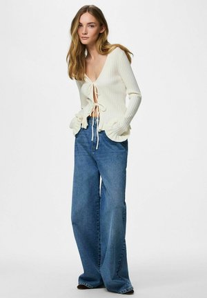 Fiatal nő fehér, hosszú ujjú, elöl megkötős bordás pulóverben és bő szabású kék farmernadrágban, egyszínű háttér előtt állva.