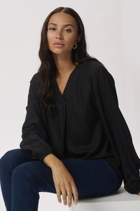Black, textured blouse with a V-neckline and billowing sleeves, paired with dark blue jeans. Model seated on a white surface, neutral background.