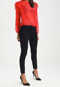 Blusa de encaje rojo con mangas con volantes, combinada con jeans ajustados negros y tacones negros de punta. El conjunto presenta un diseño texturizado.