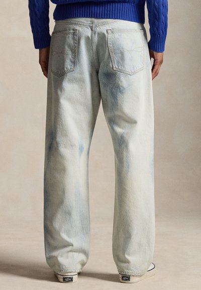Personne portant un jean clair, coupe large avec des poches arrière et un pull bleu côtelé, debout devant un fond neutre.