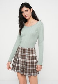 Ανοιχτό πράσινο ριμπ μακρυμάνικο τοπ σε συνδυασμό με πλισέ καρό φούστα σε καφέ και λευκό, παρουσιάζοντας αντιθετικές υφές και μοτίβα.