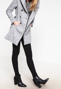 Kvinna som bär en grå dubbelknäppt kappa, svarta skinny jeans, svarta lågskor, står med händerna i fickorna mot en vit bakgrund.