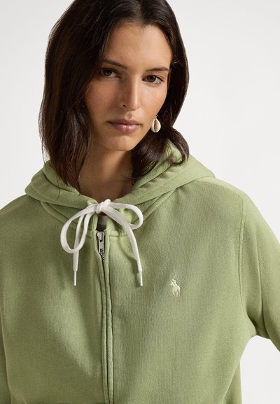 Grønn hettejakke med glidelås i mykt materiale med snøresnøring i hetten, med hvit logo på brystet og kontrasterende hvit snøring.