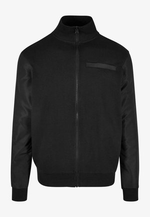 Melnā zip-up jaka ar augstu apkakli, ribotām piedurknēm un gludu tekstūru. Iekļauta maza kabata augšējā kreisajā pusē.