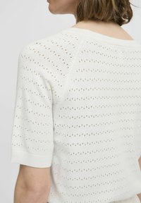 Witte trui met korte mouwen en een losse brei-stijl, voorzien van kleine ronde gaten die in horizontale lijnen zijn gerangschikt en een ronde halslijn.