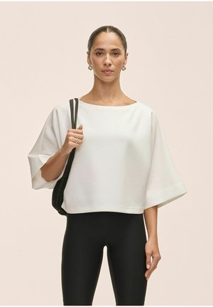 Top corto blanco con mangas amplias, hecho de una tela suave. Combinado con leggings negros y un bolso negro de hombro. Diseño simple y moderno.
