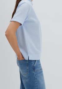 Top azzurro chiaro a maniche corte con un design a trama testurizzata tipo waffle, con taglio dritto e spacchi laterali. Abbinato a jeans in denim blu.