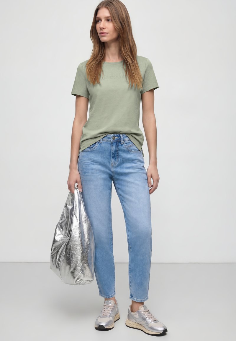 Mujer con camiseta de manga corta verde claro, vaqueros azul claro, zapatillas plateadas, sosteniendo un gran bolso tote plateado, posando contra un fondo liso.