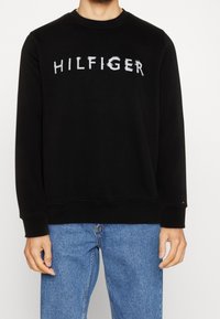 Černá mikina s potiskem slova "HILFIGER" v opotřebovaném bílém písmu. Kulatý výstřih, dlouhé rukávy, žebrované manžety a spodní lem, vyrobená z měkké látky.