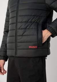 Zwarte pufferjack met een horizontaal gewatteerd ontwerp, rits aan de voorkant, elastische manchetten en een rood "HUGO" logo aan de rechteronderzijde.