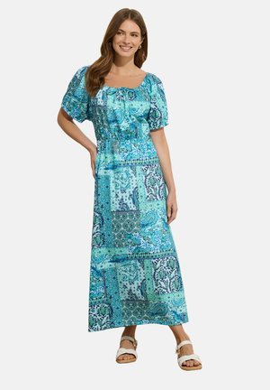 Uśmiechnięta kobieta o długich brązowych włosach, ubrana w turkusową sukienkę maxi w wzory z bufkami na rękawach i białe sandały, stojąca z jedną dłonią na biodrze.