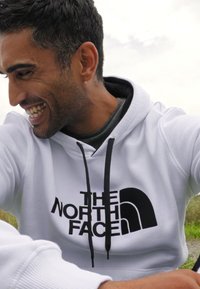 Hvit hettegenser med en svart "The North Face"-logo, med snortrukk og en myk tekstur, båret av en smilende mann i en naturlig setting.