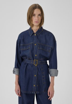 Woman wearing dark blue belted denim jacket with rolled-up sleeves and matching denim pants, standing against plain gray background.