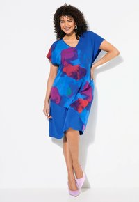 Modrá šaty s barevným abstraktním vzorem v červené a fialové. Měkký materiál, volný střih, krátké rukávy a asymetrický lem. Noseny s růžovými podpatky.