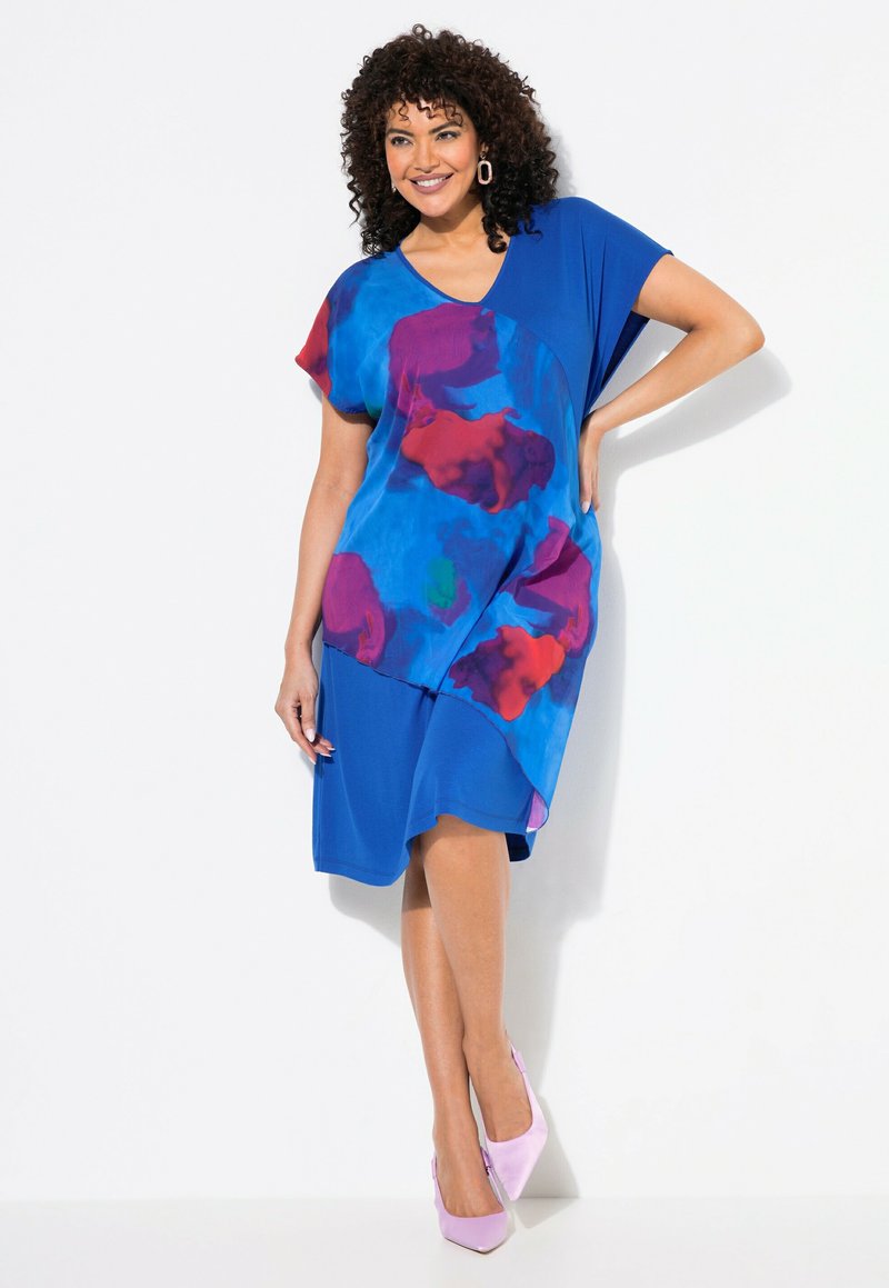 Modrá šaty s barevným abstraktním vzorem v červené a fialové. Měkký materiál, volný střih, krátké rukávy a asymetrický lem. Noseny s růžovými podpatky.