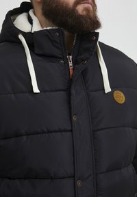 Polstret jakke i mørkeblå med hætte foret med fleece, der har en lynlåslukning fortil, dekoreret med brune knapper og et emblem patch.