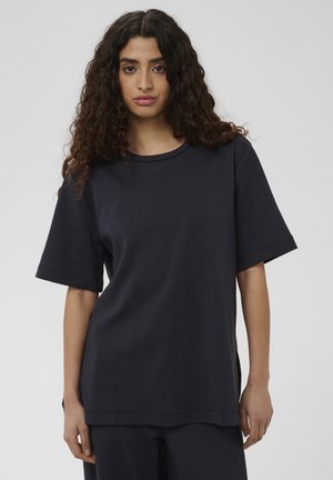 Jonge vrouw met lang krullend haar, dragend een loszittend donkerblauw T-shirt met korte mouwen en bijpassende broek, staand tegen een egale achtergrond.