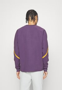 Sudadera morada con cuello redondo, puños de canalé y rayas amarillas destacadas en las mangas; fabricada con un tejido suave y texturizado.