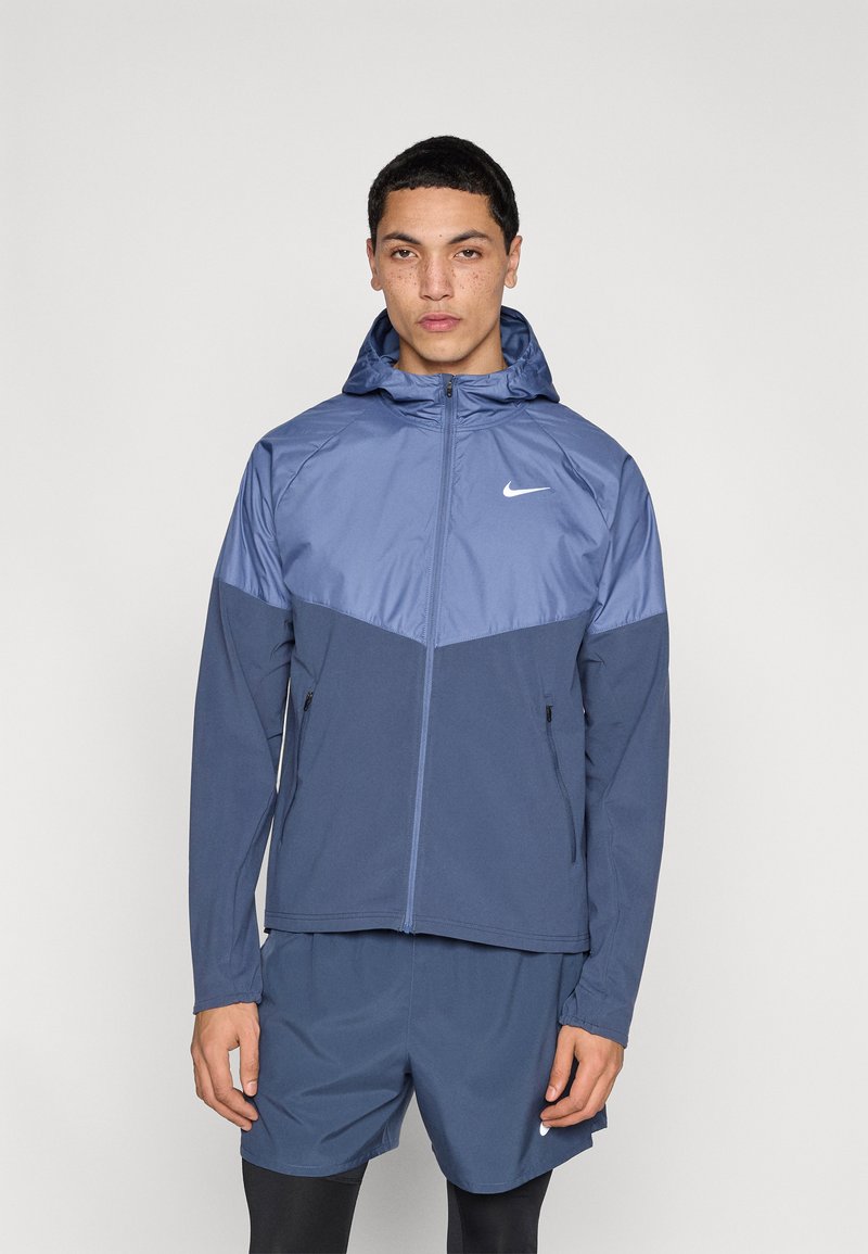 Μπουφάν με κουκούλα της Nike σε μπλε αποχρώσεις, με φερμουάρ μπροστά, ελαστικά μανίκια και δύο πλαϊνές τσέπες. Λείο υλικό με σχέδιο color-block.