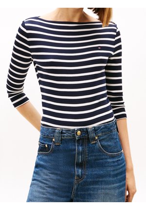 Femme portant un chemisier à manches longues rayé bleu marine et blanc, associé à un jean bleu taille haute, sur fond blanc.