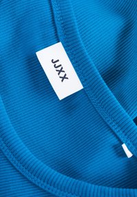 Tecido azul com nervuras e uma textura suave. Apresenta um decote curvo e uma etiqueta branca com a inscrição "JJXX."