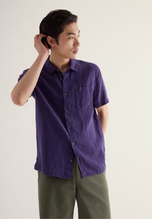 Mees, kes kannab lühikeste varrukatega lilla nööpidega särk ja oliivrohelisi pükse, vaatab kõrvale ning puudutab üht käega oma juukseid.