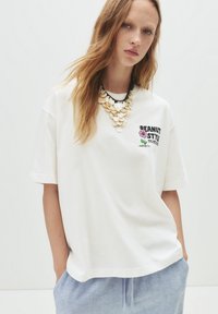Hvid bomulds t-shirt med korte ærmer, der har et grafisk print med teksten "PEANUT STYLE" og en blomst. Modellen bærer en stor halskæde.