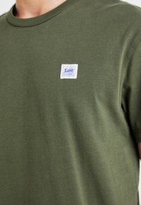 Olijf groene katoenen t-shirt, ronde hals, korte mouwen. Heeft een blauw en witte rechthoekige Lee Workwear label op de borst.