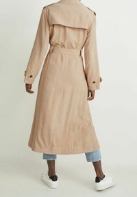 Beżowy trencz o luźnym kroju, z długimi rękawami, paskiem w talii i klapą z tyłu, zestawiony z obcisłymi dżinsami i białymi sneakersami.